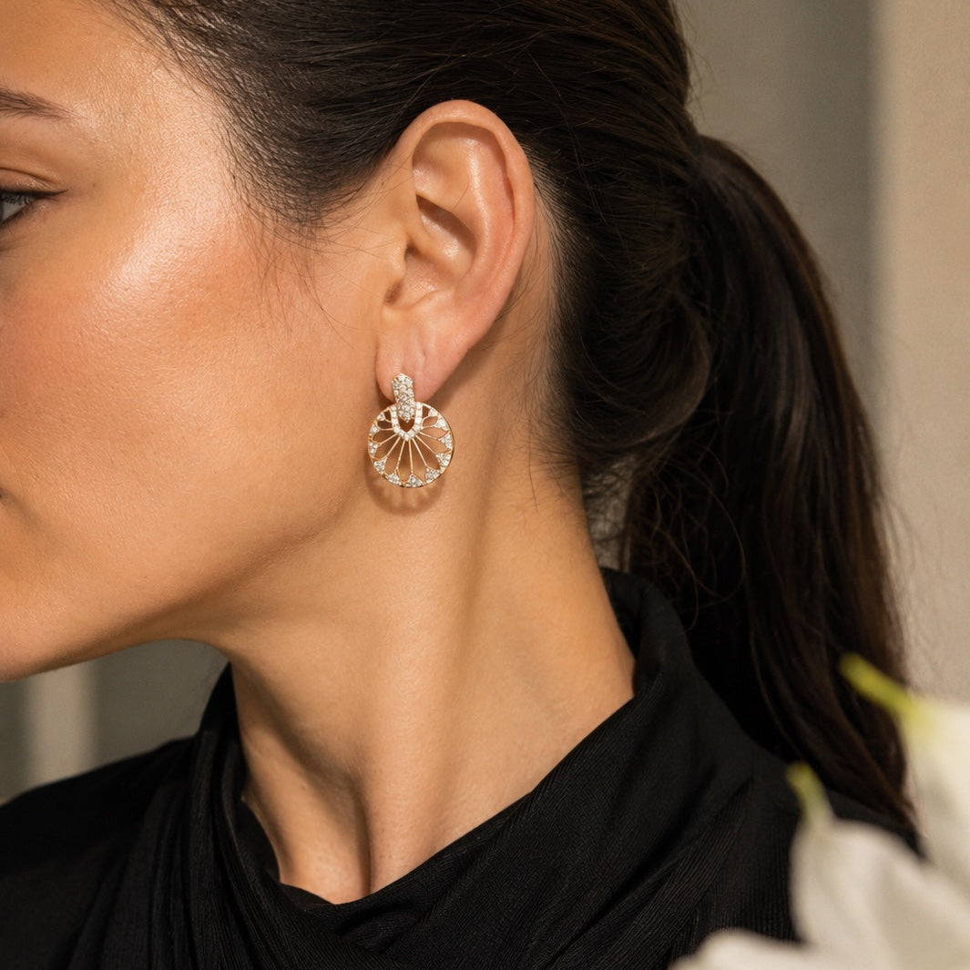 A close-up side profile of a woman with dark hair tied back, wearing a delicate gold-toned earring with a sunburst or fan-like design and small clear stones, paired with a black top.