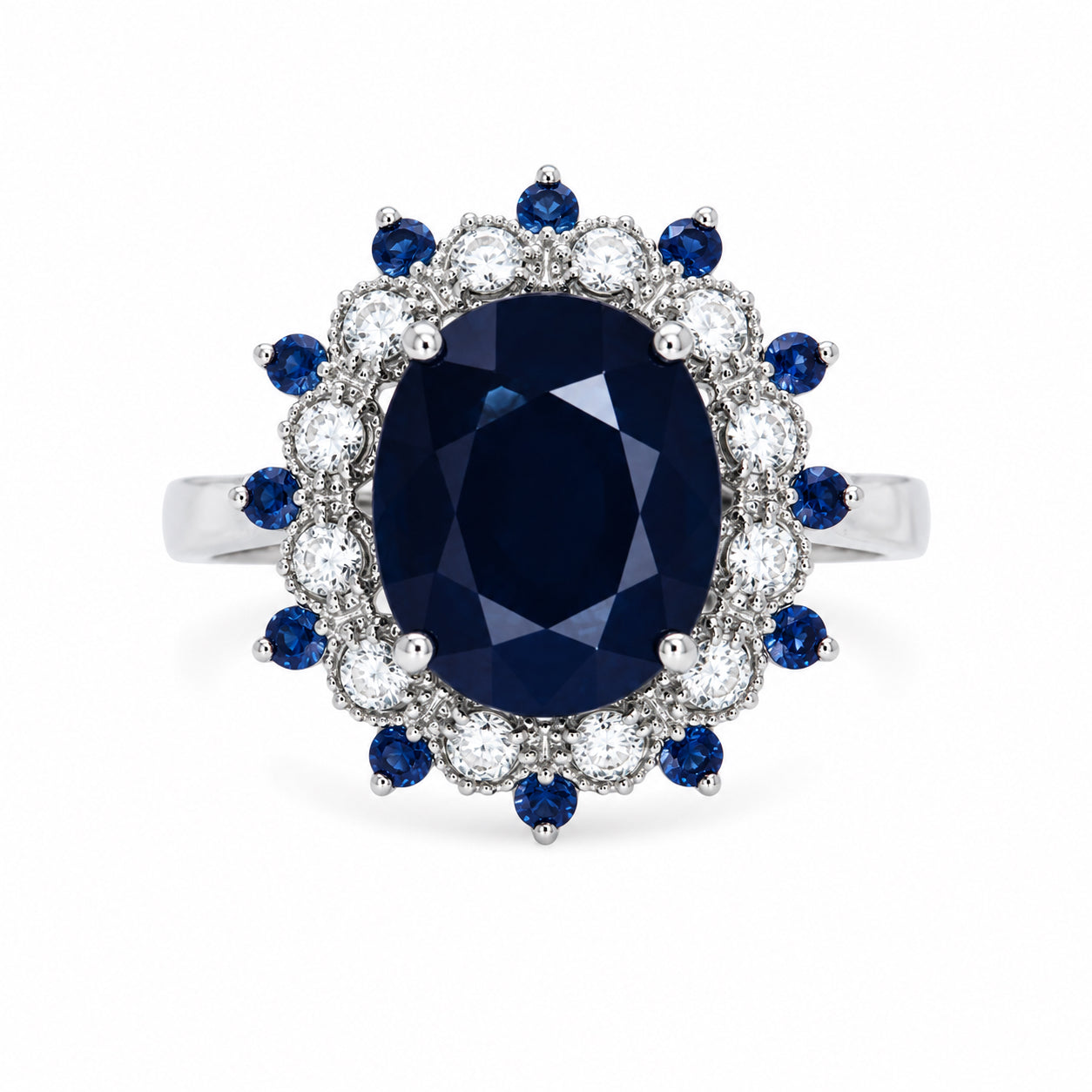 An oval-cut sapphire cluster ring with a halo of small blue sapphires and round clear diamonds on a polished silver-colored band, set against a plain white background.