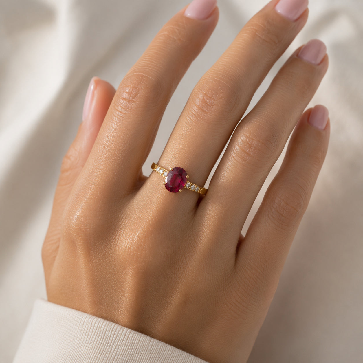 A gold ring with an oval ruby gemstone and small side stones, worn on a hand against a soft white fabric background.